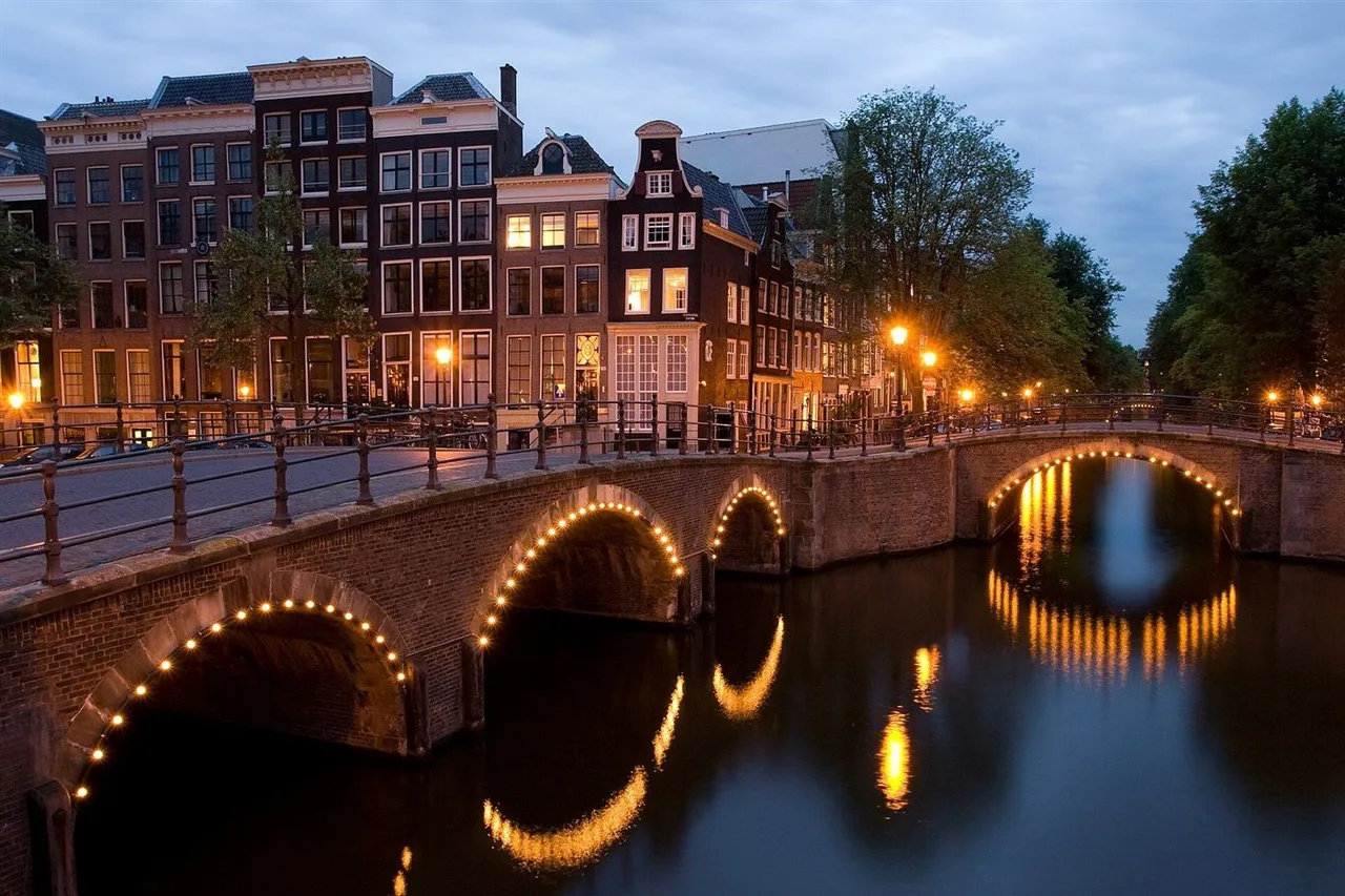 Brick bridges with lit arches crossing a canal at dusk, with tall gabled brick houses and trees along the water.