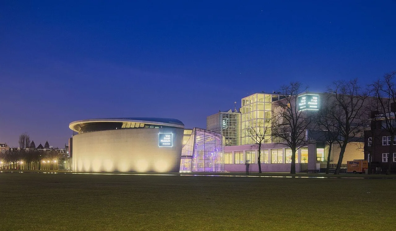 A curved pale building and a glass-fronted entrance lit at dusk beside a dark lawn, with illuminated Van Gogh Museum signs.
