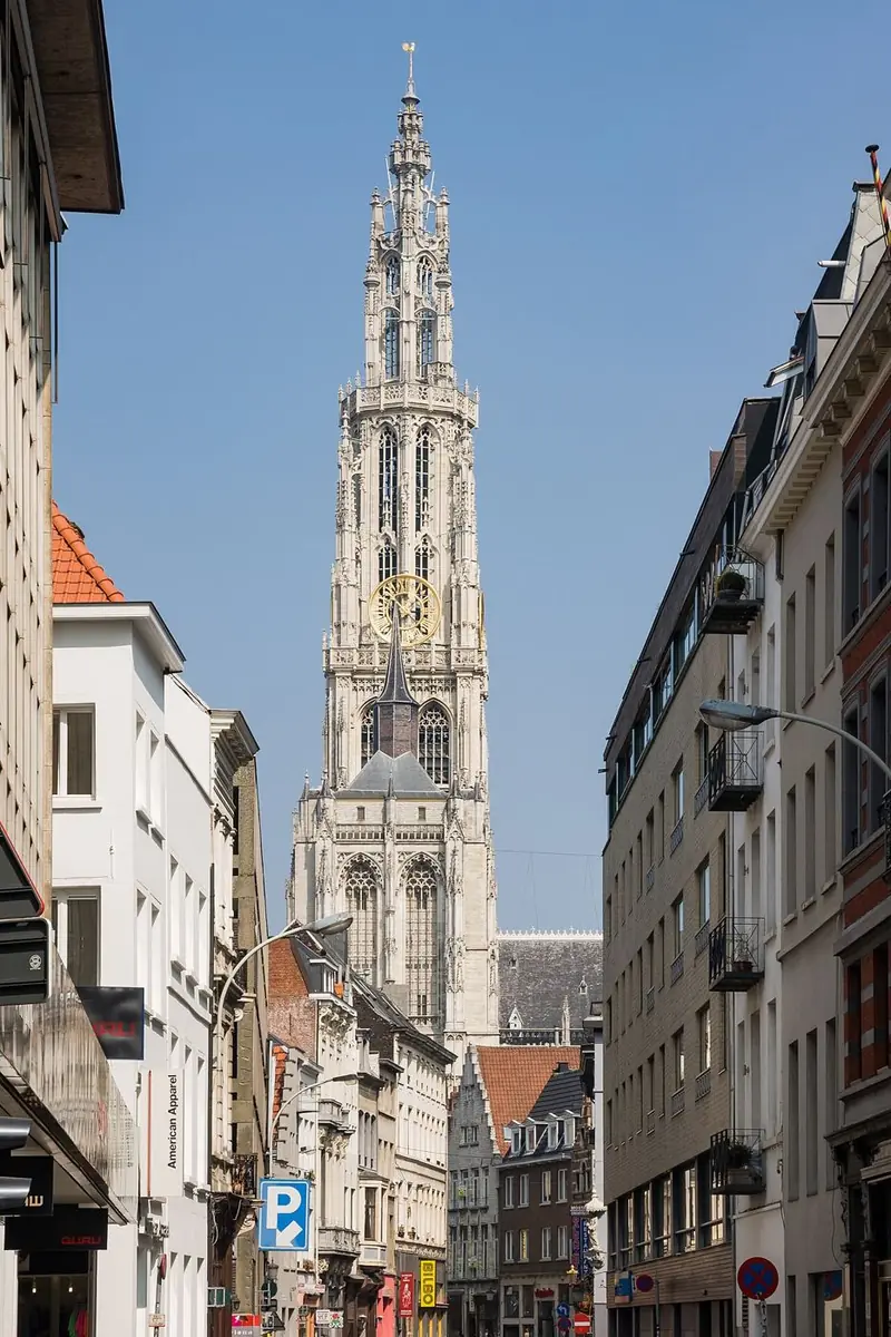 The north tower of Antwerp's Cathedral of Our Lady rising above a narrow street of shops and townhouses, with a gold clock face near the top and a tall openwork spire.