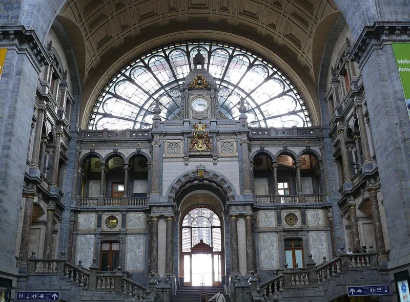 The grand entrance hall of Antwerp Central Station, with a marble-clad double staircase, a large arched glass window above a clock and gilded coats of arms.