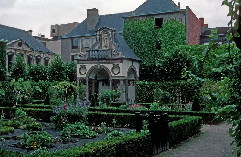 A garden of box-edged beds with flowers in front of a small stone pavilion with an arched portico and a statue in a niche, with ivy-covered walls behind.