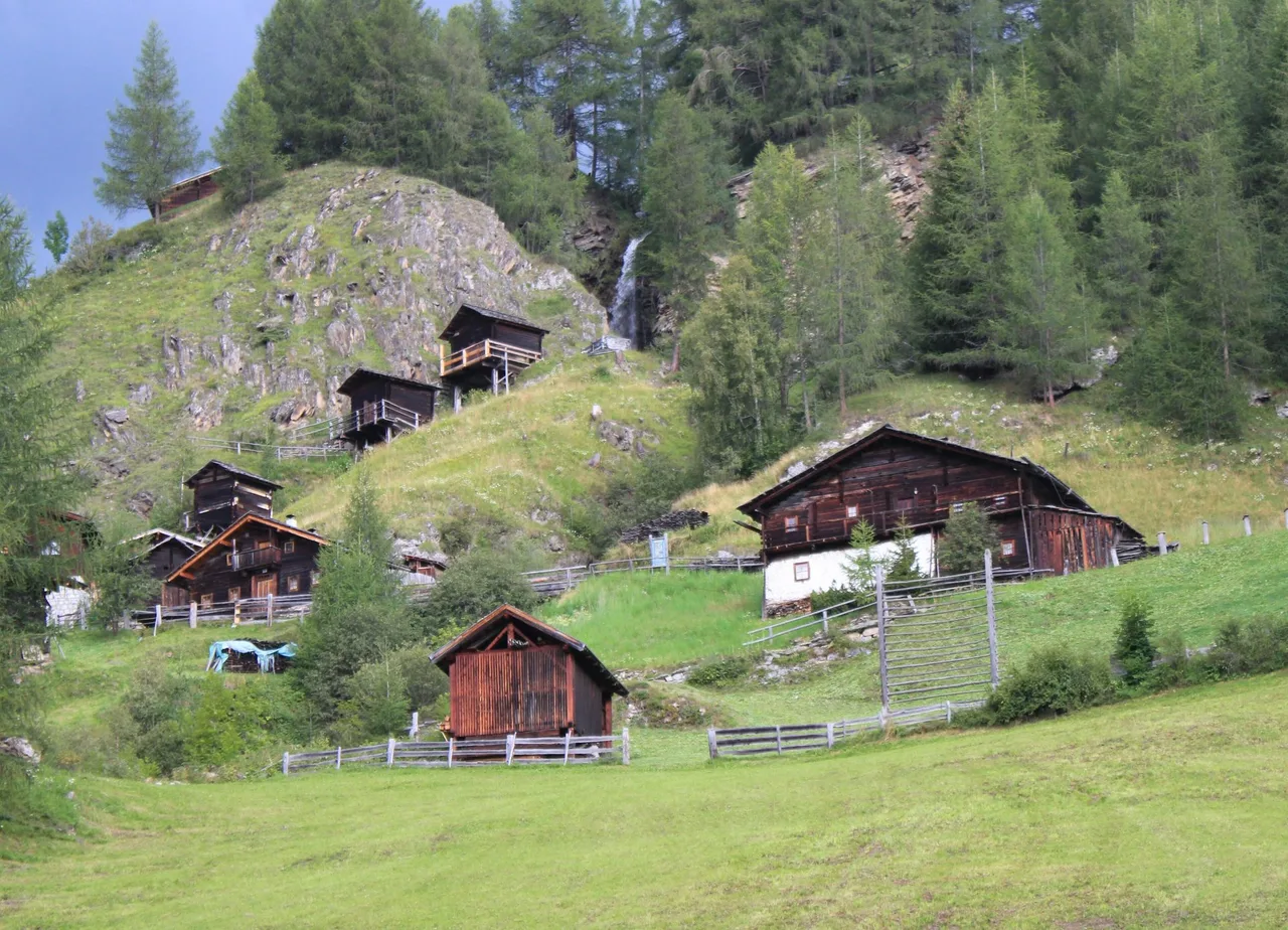 Dark wooden mills and farm buildings on stilts on a steep green slope in Apriach, with a small stream and forest above