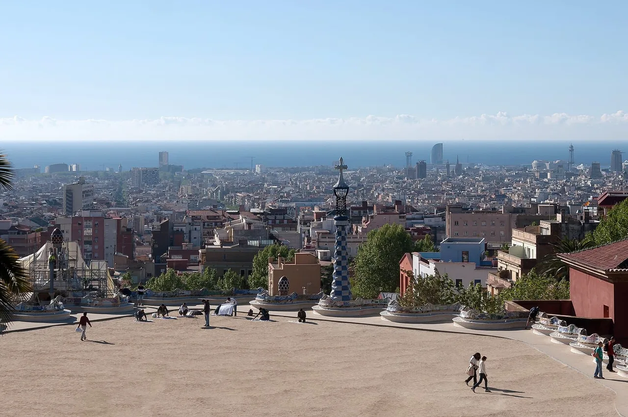 The open terrace of Park Güell with a tiled column and curved benches, and the roofs of Barcelona stretching to the sea on the horizon.