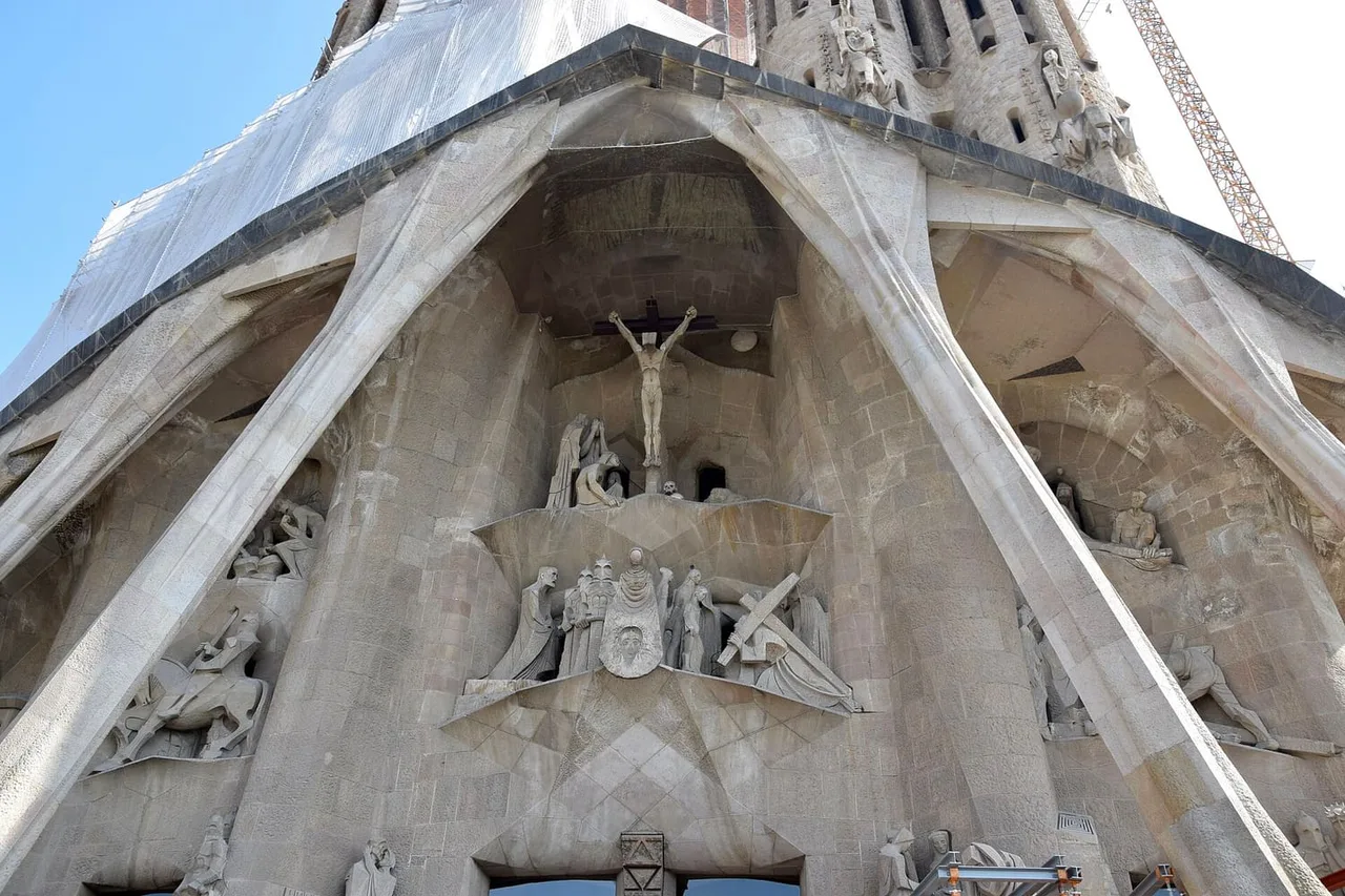 Sculpted figures and a crucifix under the sloping stone buttresses of the Sagrada Família Passion façade, with scaffolding and a crane at the edge of the frame.