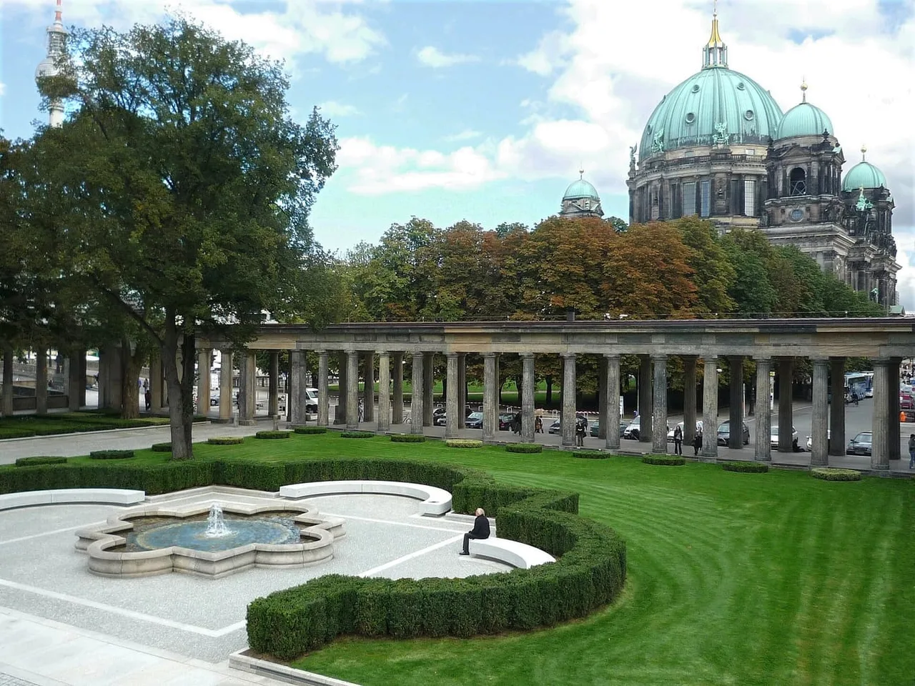 Garden court with a fountain and a stone colonnade, with the green copper dome of Berlin Cathedral and the television tower behind.