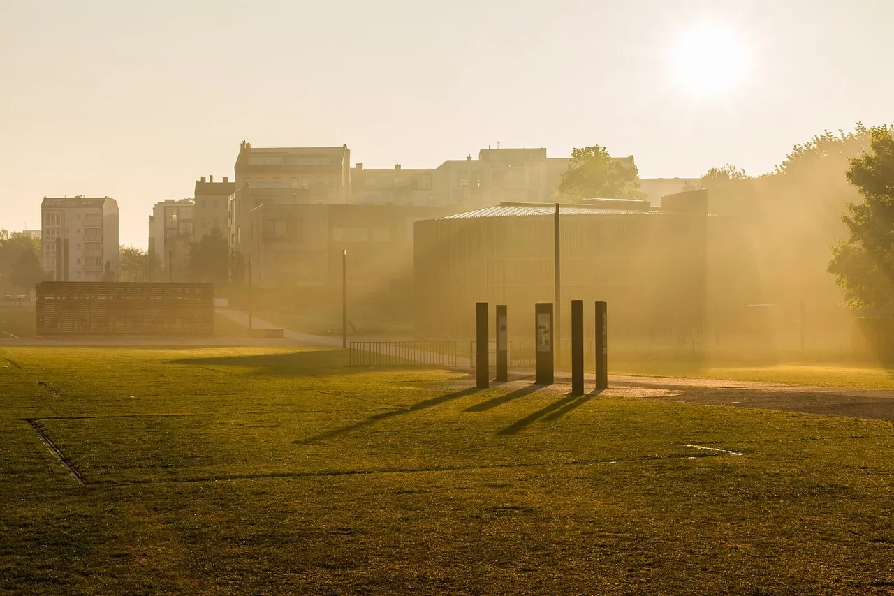 Information stelae on a grass field in morning haze, with a rounded dark building and apartment blocks behind.