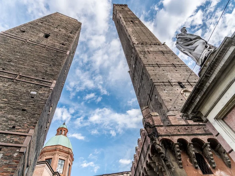 The two leaning brick towers of Bologna seen from below against a blue and white sky, with a green church dome and a statue between them.