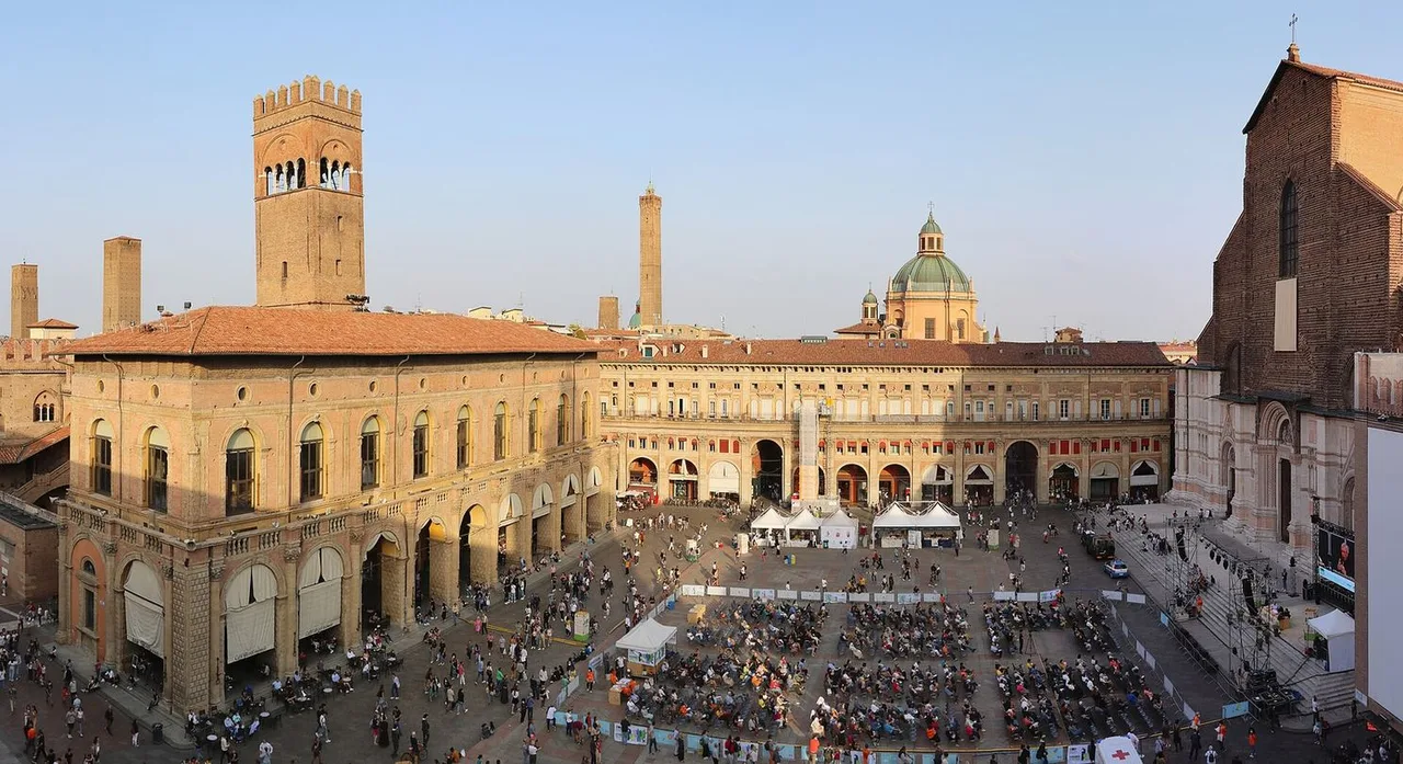 Piazza Maggiore seen from above with a crowd gathered in the square, a medieval palace tower at left, the brick side of a large church at right and a green dome behind.