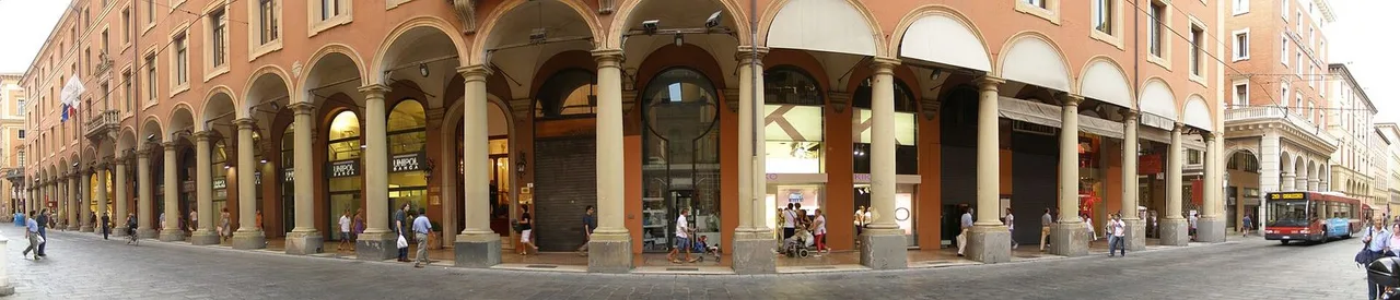 A long covered walkway under repeated round arches along Via Indipendenza, with shop fronts, a city bus and a few pedestrians in the distance.
