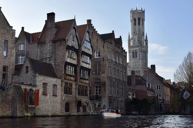 A tourist boat on a canal in Bruges beside tall brick and timber-fronted houses, with the Belfry tower rising against a pale blue sky and a swan in the foreground.