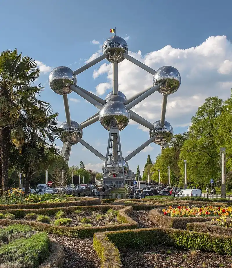 The Atomium, nine large metallic spheres joined by tubes, seen from a garden with clipped hedges and tulips under a blue sky with white clouds.