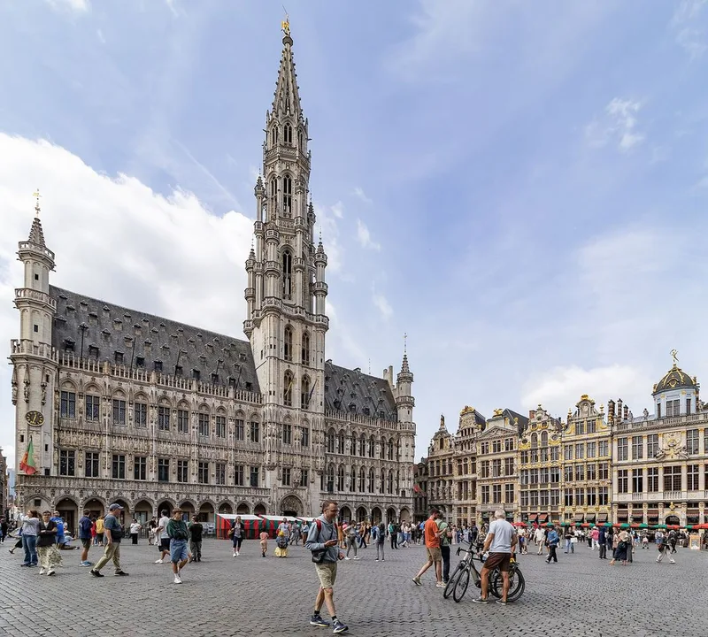 Brussels Town Hall with its tall Gothic spire and a row of gilded Baroque guild houses facing a cobbled square with strolling visitors and a parked bicycle.