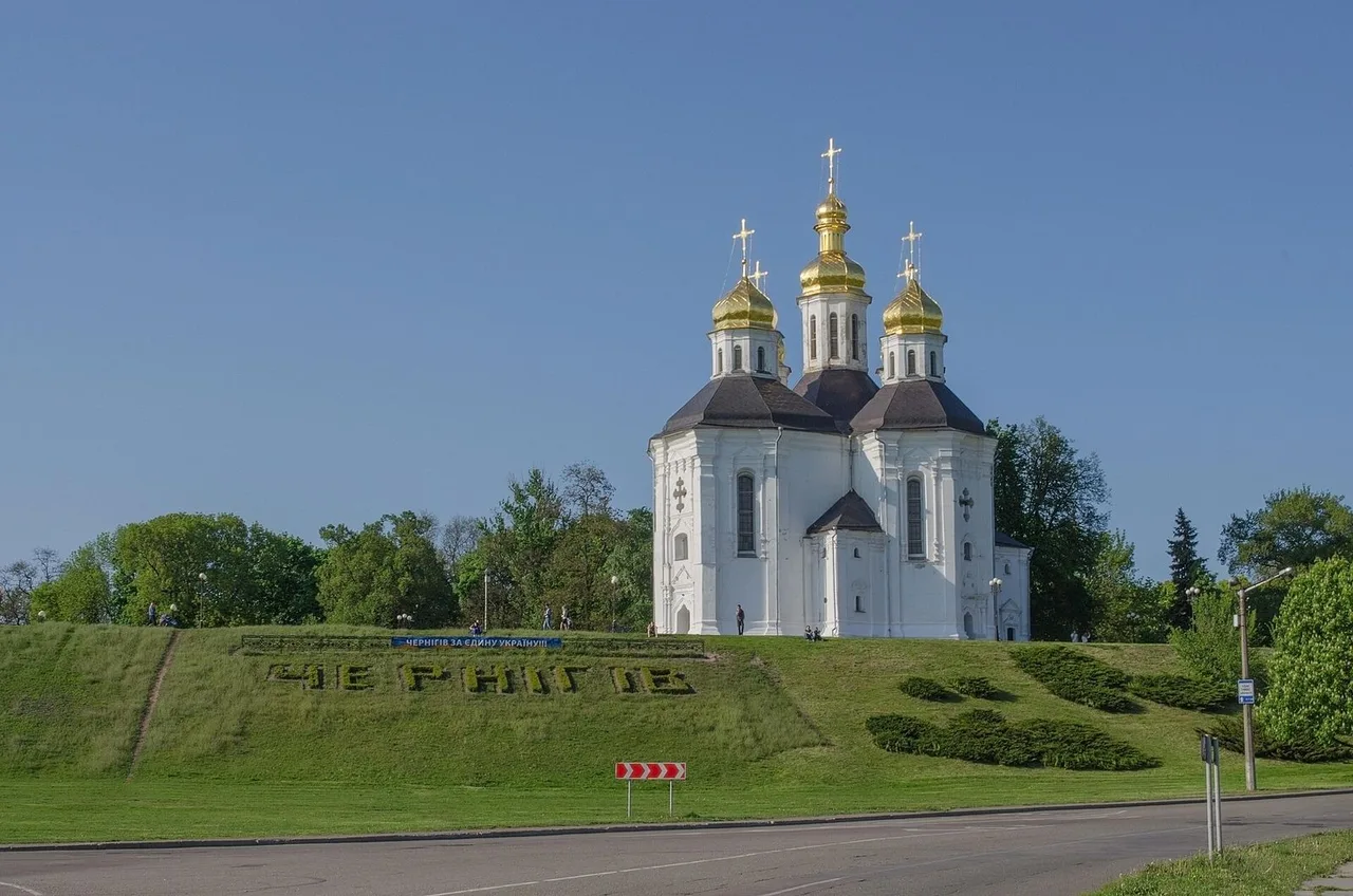 A white Baroque church with gilded domes on a grassy slope with clipped bushes, under a blue sky.