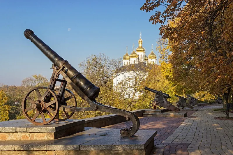 Old iron cannons on wheeled carriages on a paved rampart, with a white church with gilded domes among autumn trees behind.