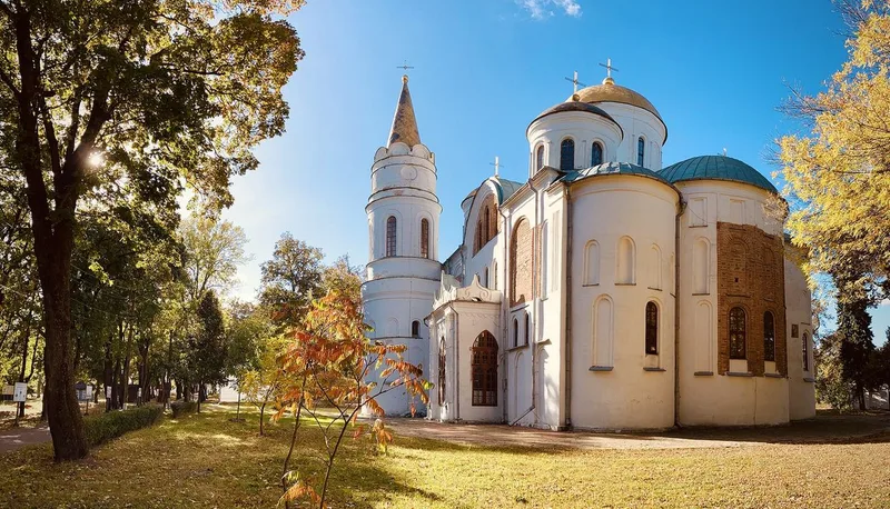 The white Transfiguration Cathedral in Dytynets Park with a tall tower and domes among trees in autumn colours.