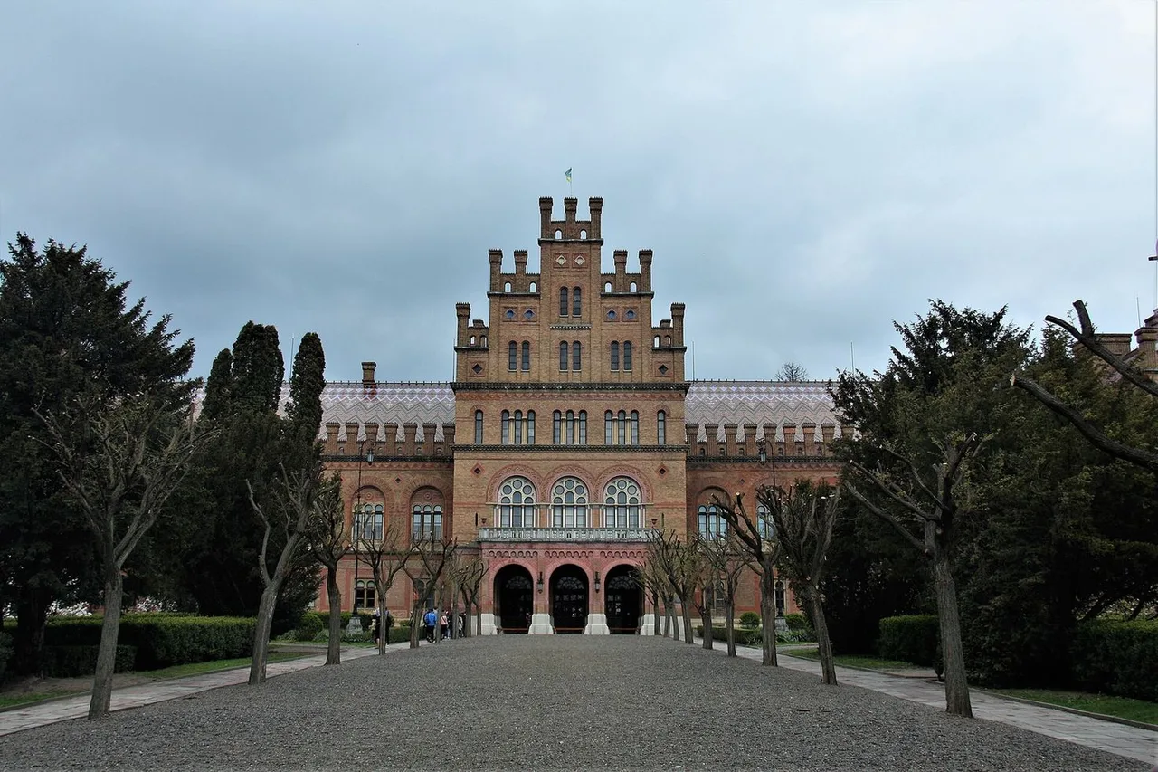 The main facade of the red-brick university buildings along a gravel path between trees, with a stepped central gable and three arched doorways.