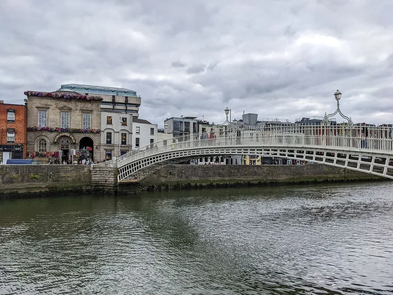 A white cast-iron footbridge arching over the River Liffey, with terraced buildings along the quay under a cloudy sky.