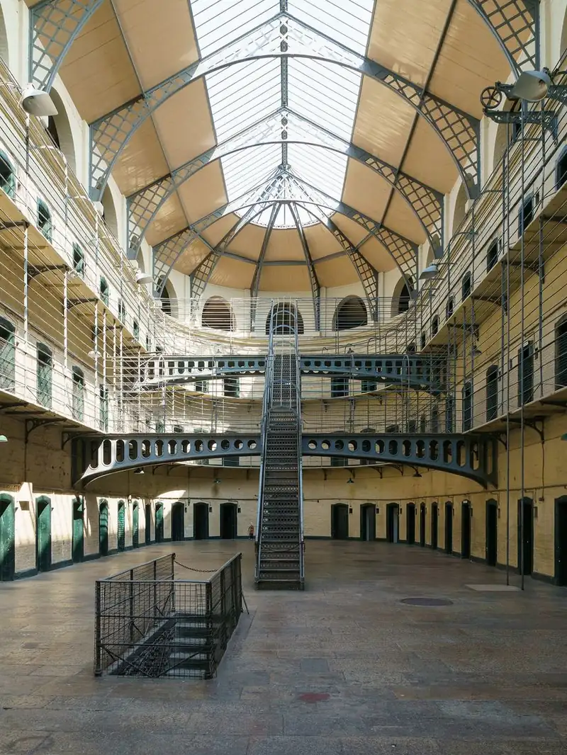 A tall prison hall with a glass roof, tiers of cell doors, iron walkways and a central staircase.