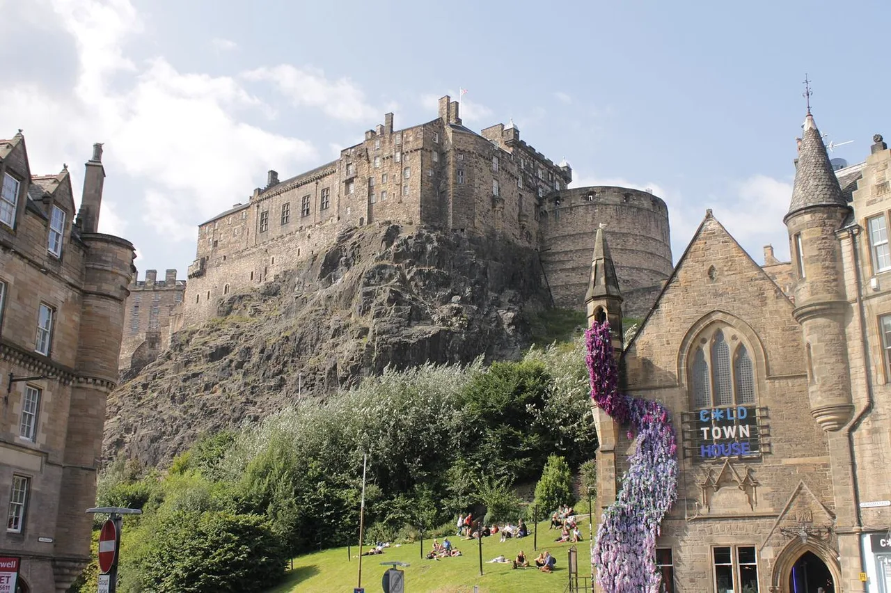 Edinburgh Castle on its rocky crag above the Grassmarket, with Victorian stone buildings in the foreground and people on the lawn below.