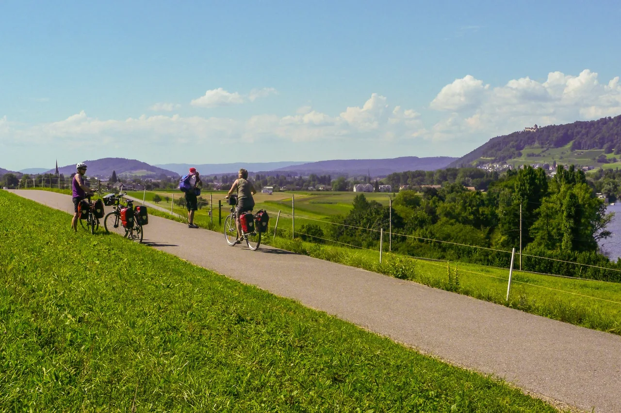 Three cyclists with panniers stop on a paved path along a grassy dike beside the Rhine, with hills and a hilltop castle in the distance.