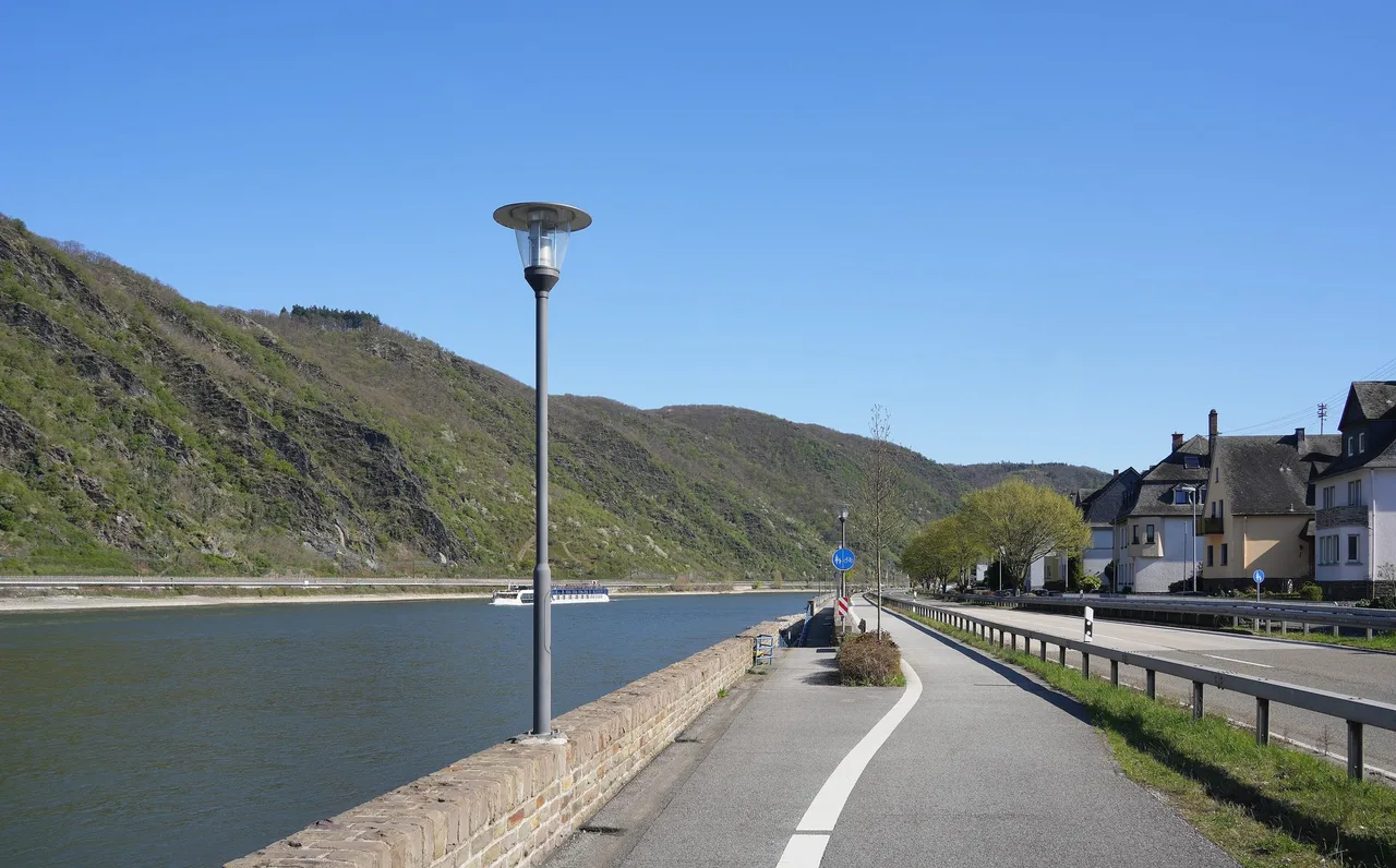 A separated cycle path with a white line runs along a stone river wall beside the Rhine, with wooded hills across the water and a road on the other side.