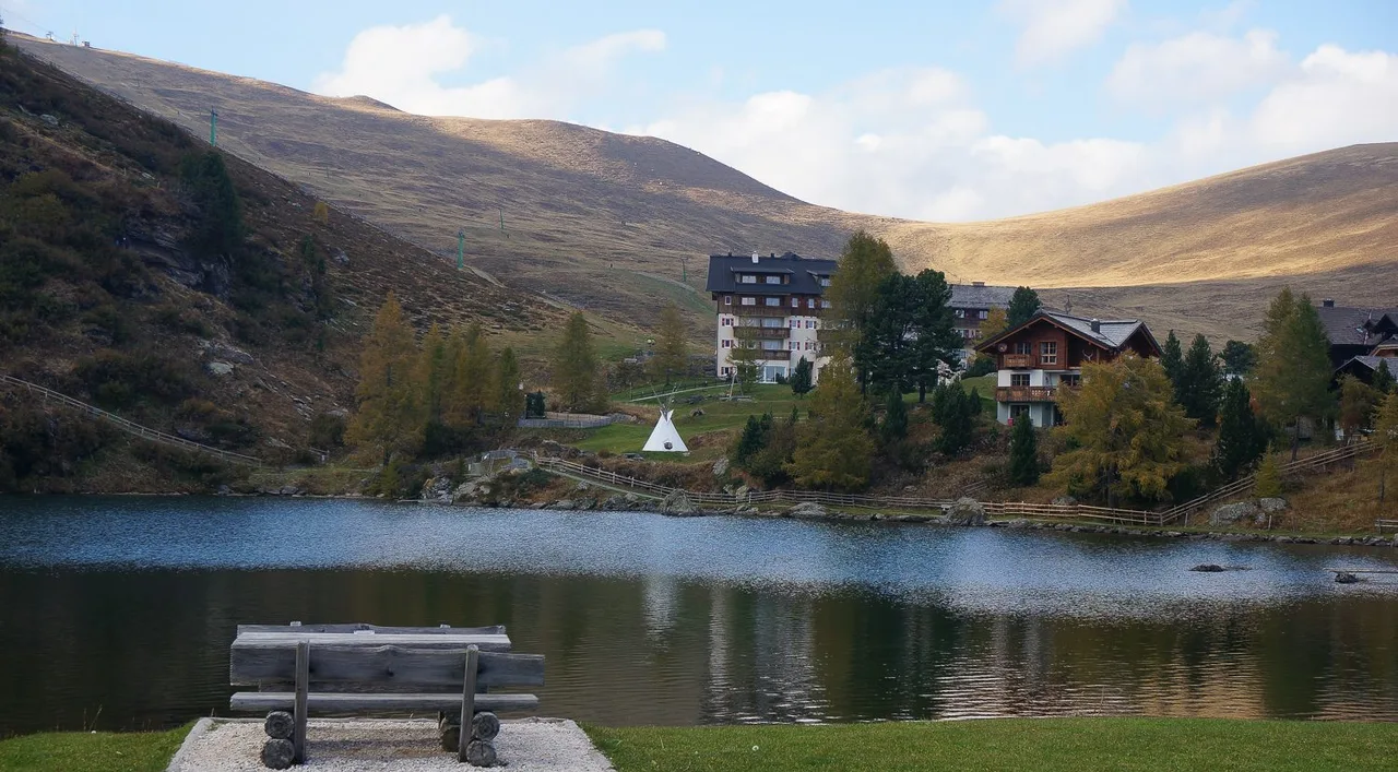 Still lake in the foreground with a bench, a hotel and wooden houses on the far shore and bare rounded hills above.