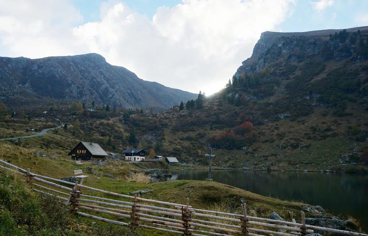 Small mountain lake with a wooden cross on its shore, a few huts and steep rocky slopes rising behind.