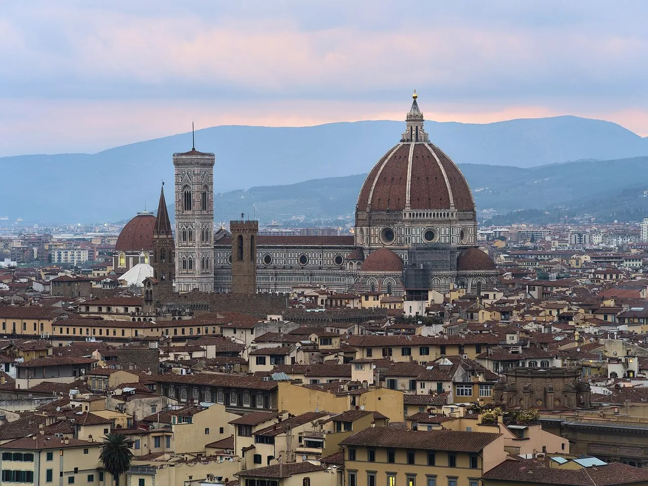 Red-tiled rooftops of Florence with the cathedral dome and the tall Bell Tower in the middle, hills rising behind under a pink evening sky.