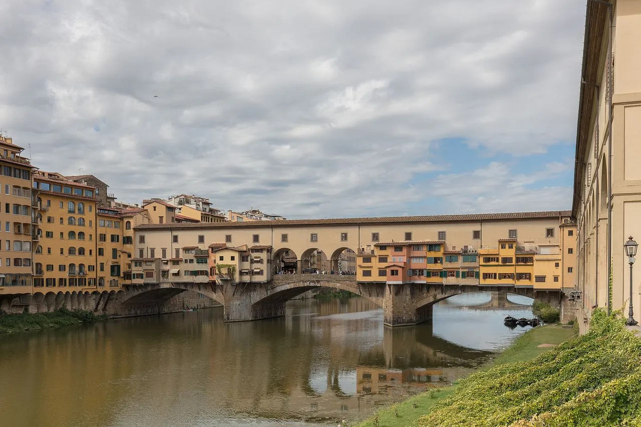 Ponte Vecchio crossing the Arno, lined with shops and houses in warm colours, with a green riverbank in the foreground.