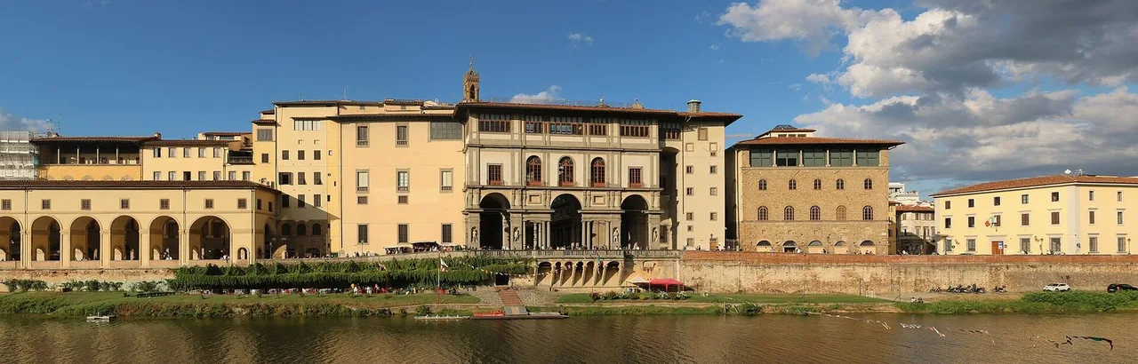 The long ochre facade of the Uffizi buildings with rows of arches beside the Arno, seen from across the river under a blue sky.