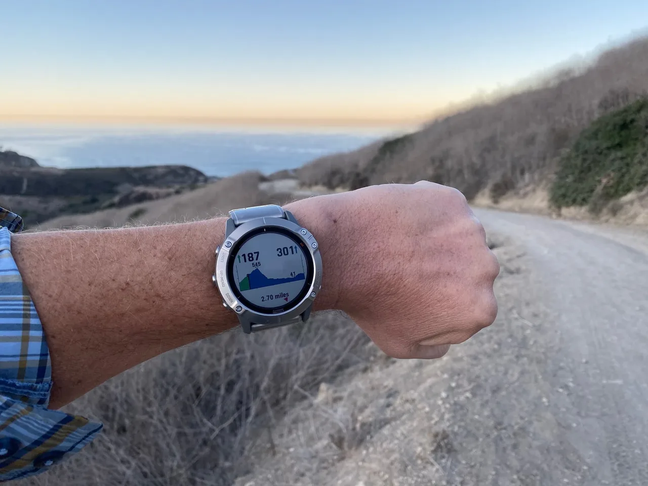 A wrist wearing a Garmin fēnix 6X Pro Solar showing an elevation profile, held out over a gravel track on a dry hillside at dusk.