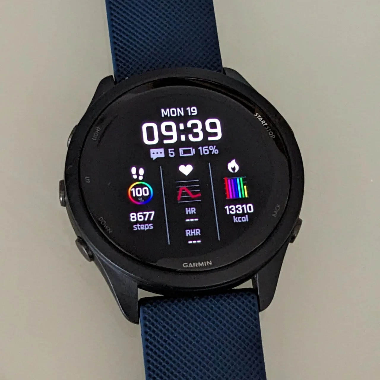 A black Garmin Forerunner 265 with a blue textured band, lying flat and showing steps, heart rate and calories on the watch face.