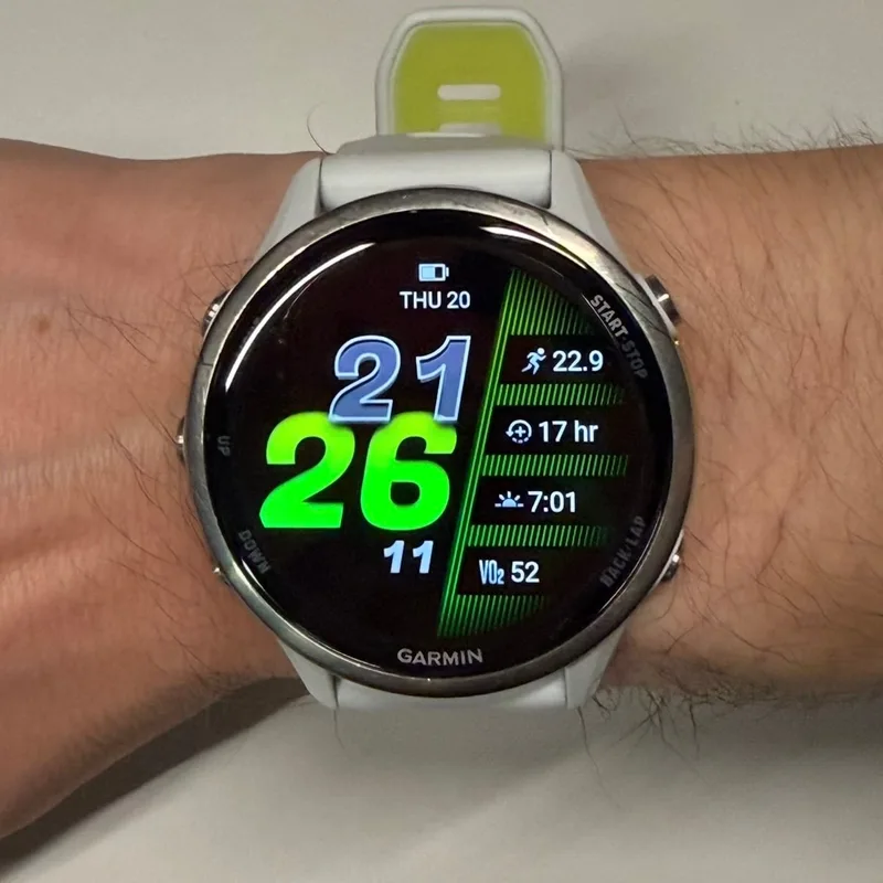 A Garmin Forerunner 970 with a white band and silver bezel on a wrist, with a green and blue digital watch face and the Start-Stop and Back-Lap button labels.
