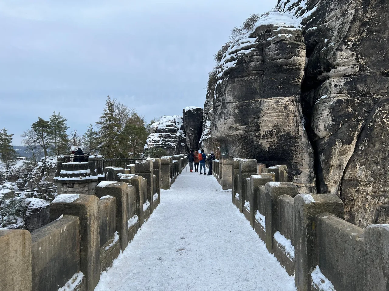 Snow-covered Bastei bridge with sandstone parapets and tall rock towers on a winter day, with a few small figures in the distance