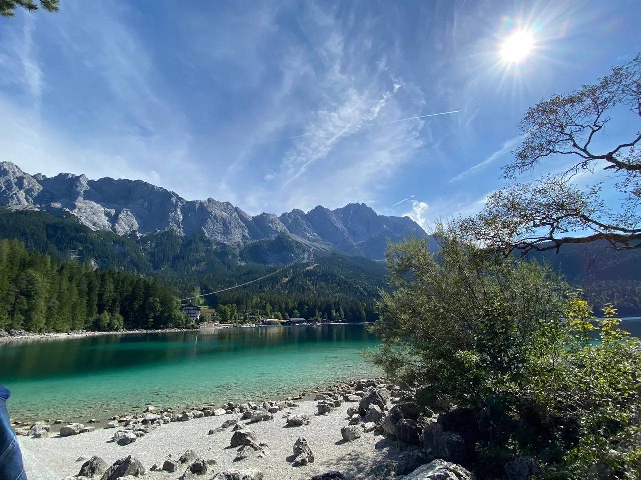 Turquoise water of the Eibsee with a pebble shore, forest and the Wetterstein mountain wall rising behind, with a cable car line crossing the slope