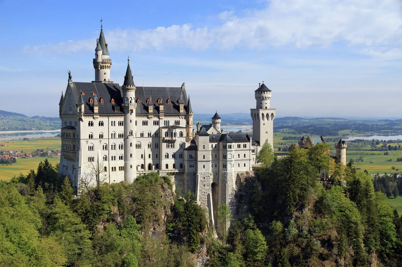 Neuschwanstein Castle on its rock, seen from the direction of the Marienbrücke, with green fields and lakes in the plain behind