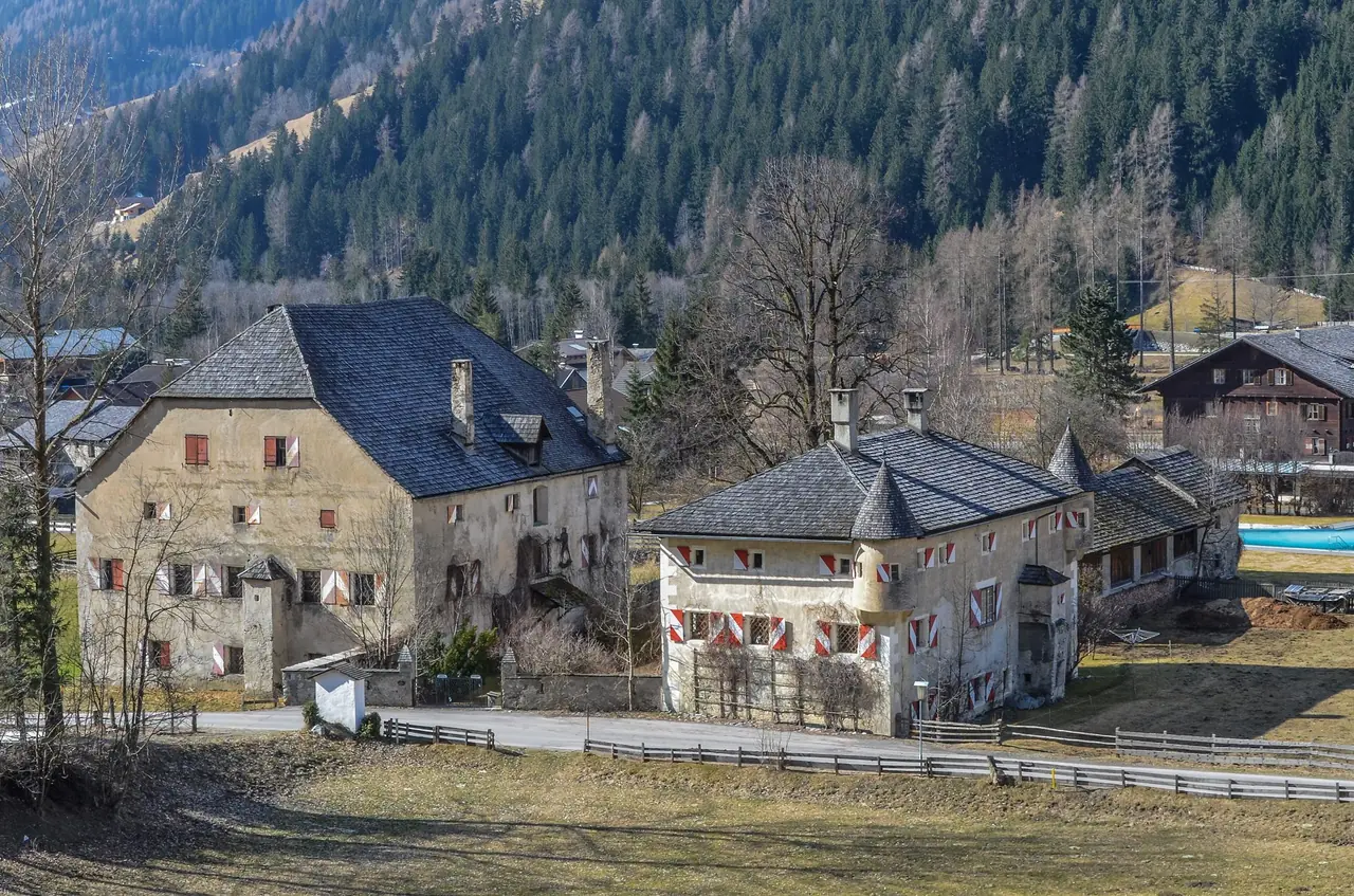 Two historic manor buildings with slate roofs and red and white shutters in Döllach, with a wooded slope behind
