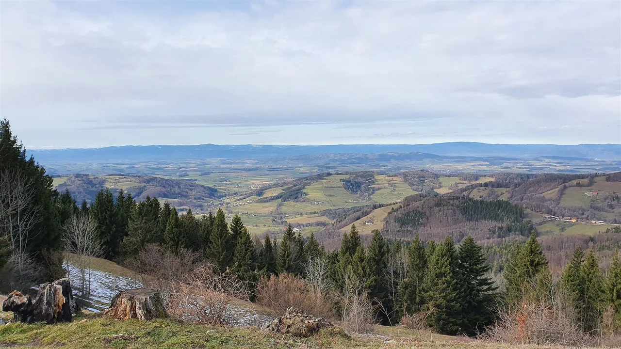 A wide view over rolling farmland and forested hills in early spring, with small patches of snow on the slope in the foreground.