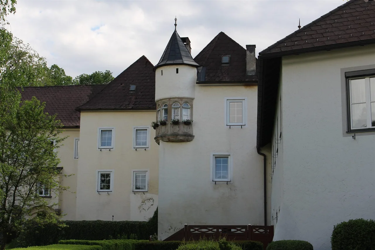 Cream-coloured castle walls with a round corner tower, a stone oriel window and dark roofs behind a clipped hedge.