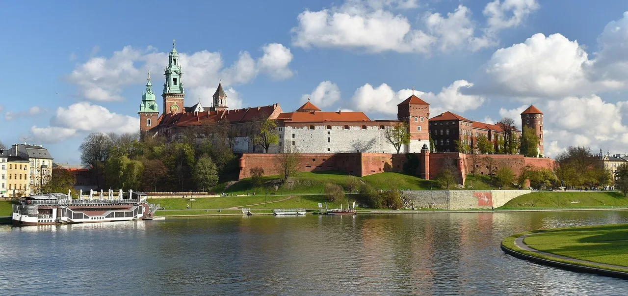 Wawel Hill with red-roofed castle buildings and a green-topped cathedral tower above grassy banks and the Vistula, with a river boat moored at left.