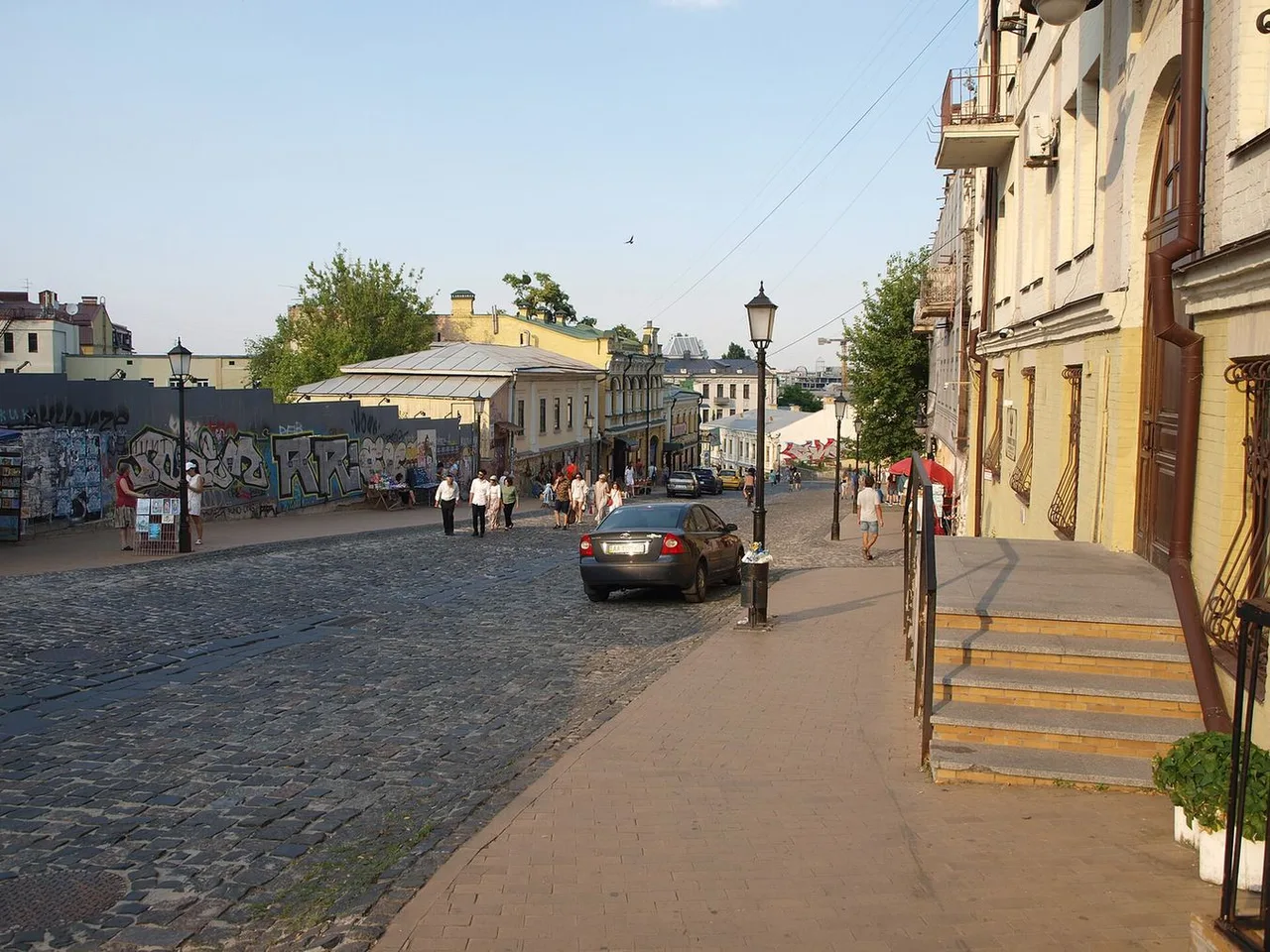 A cobbled stretch of the Andriyivskyi Descent with a car, street lamps, a graffiti wall and pedestrians in the distance.