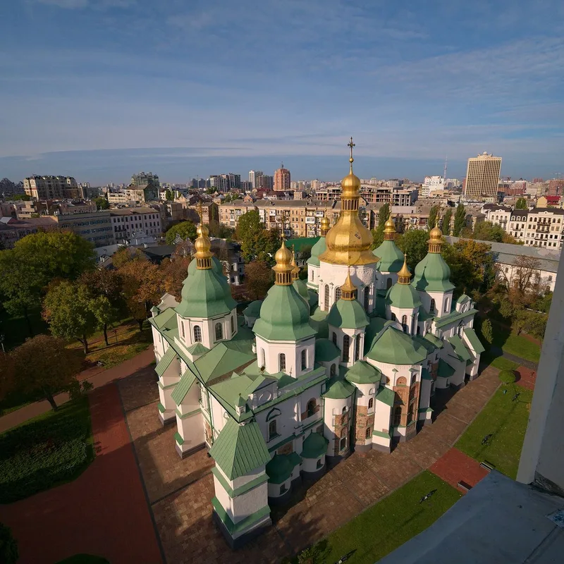 Saint Sophia Cathedral seen from above, with green roofs, white walls and gilded domes among gardens and city buildings.