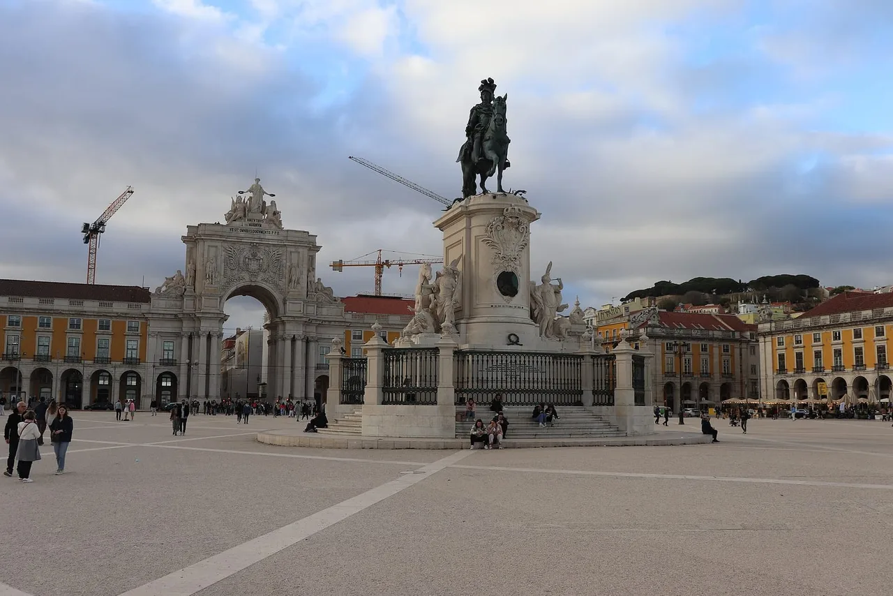 A wide paved square with an equestrian statue on a stone plinth, a triumphal arch at the far side and yellow arcaded buildings under a cloudy sky.