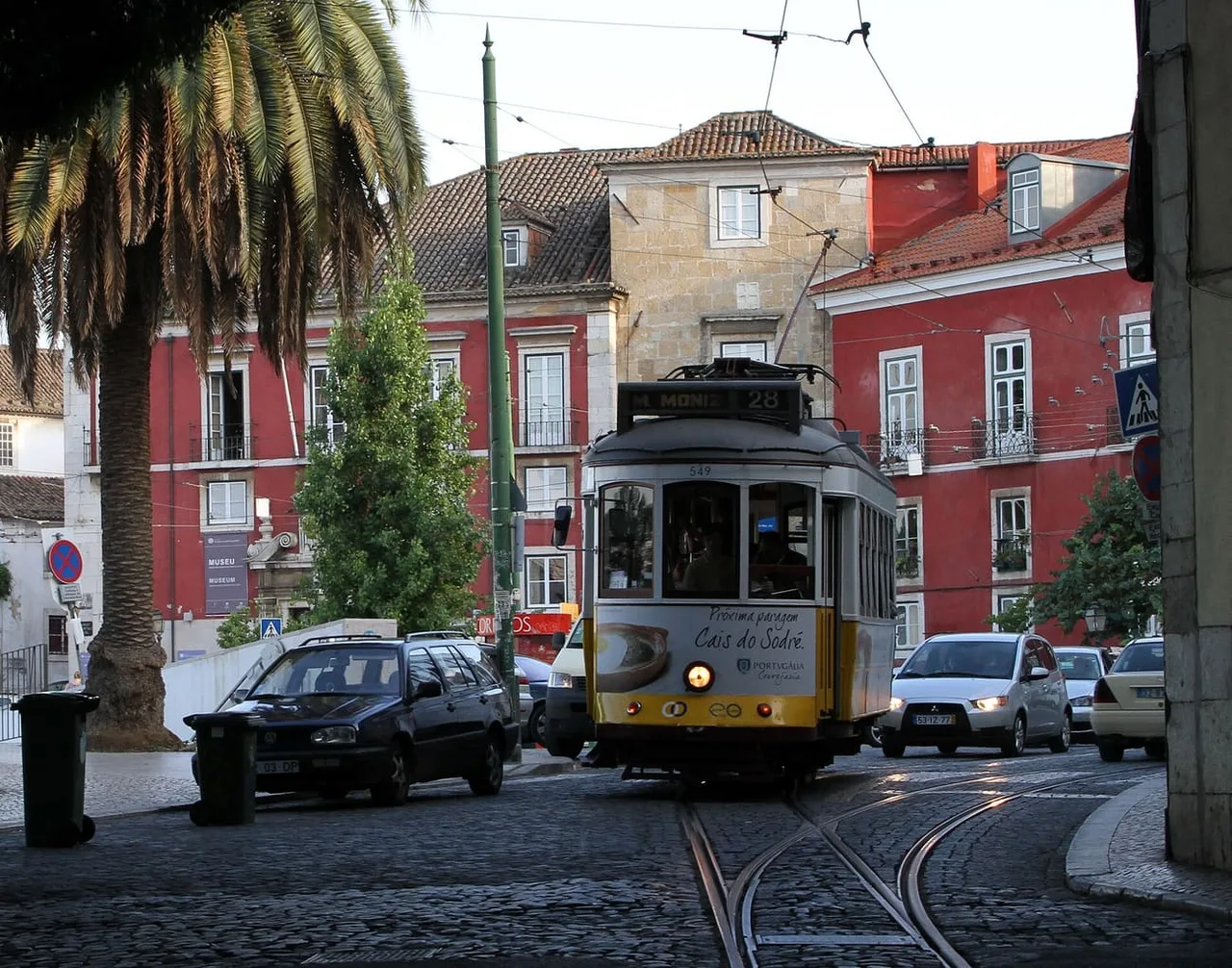 A yellow and white tram marked 28 on cobbled tracks in a narrow street of red and ochre buildings, with a palm tree at the left.