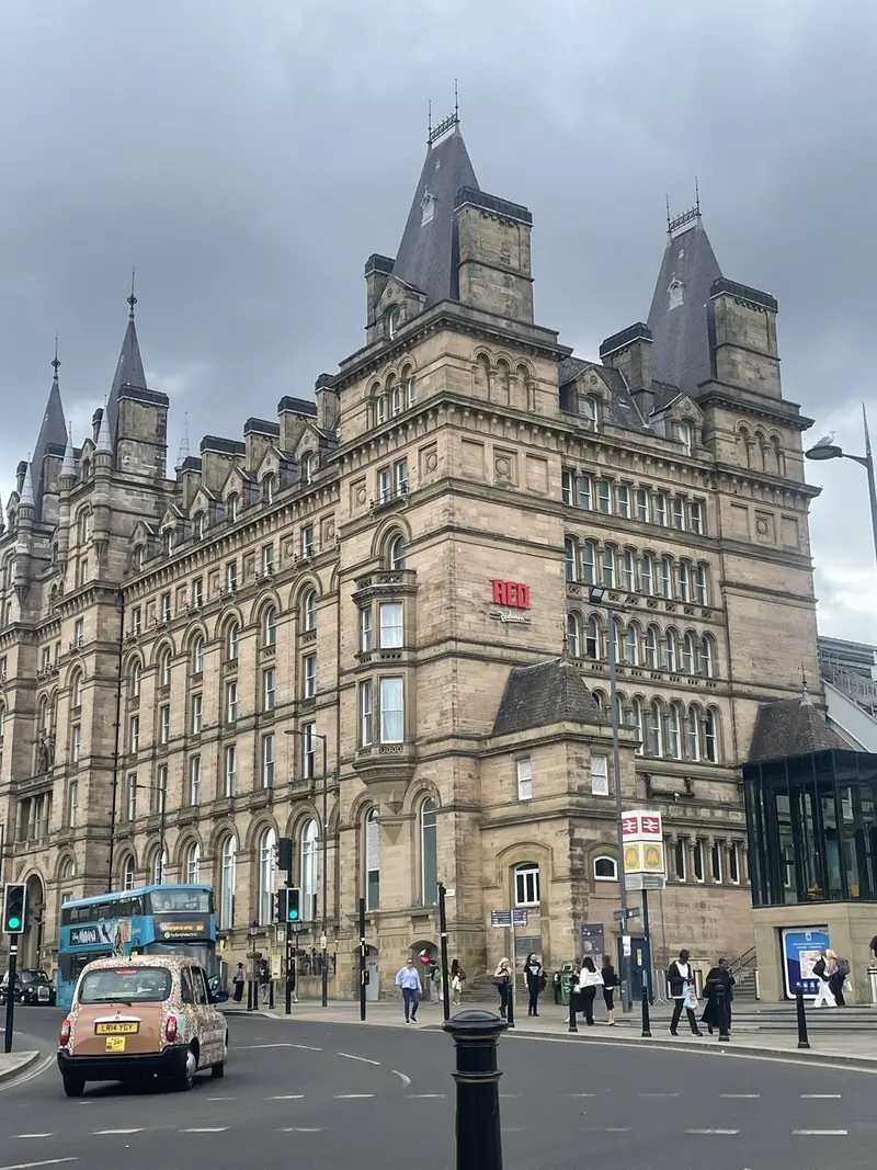 A Victorian Gothic stone building with pointed turrets and steep roofs on a street corner, with a pink taxi and a bus in front.