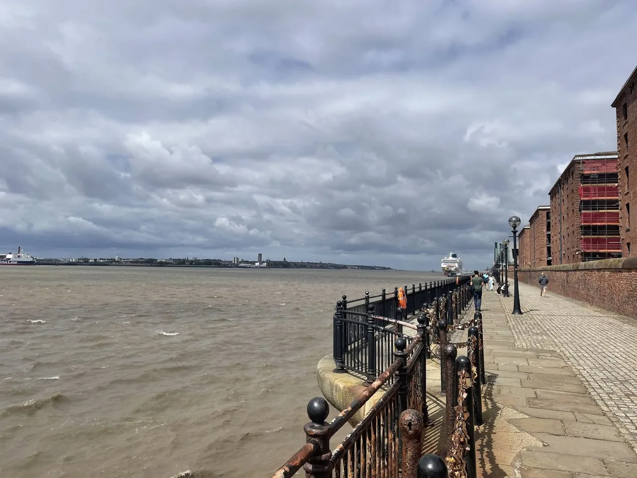 The brown River Mersey beside a railed riverside promenade, with a red-brick dock warehouse at the right under cloudy skies.