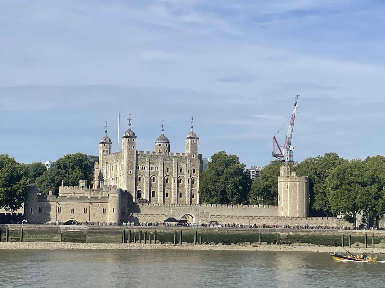 The White Tower and the stone walls of the Tower of London seen across the Thames at low tide, with trees behind.