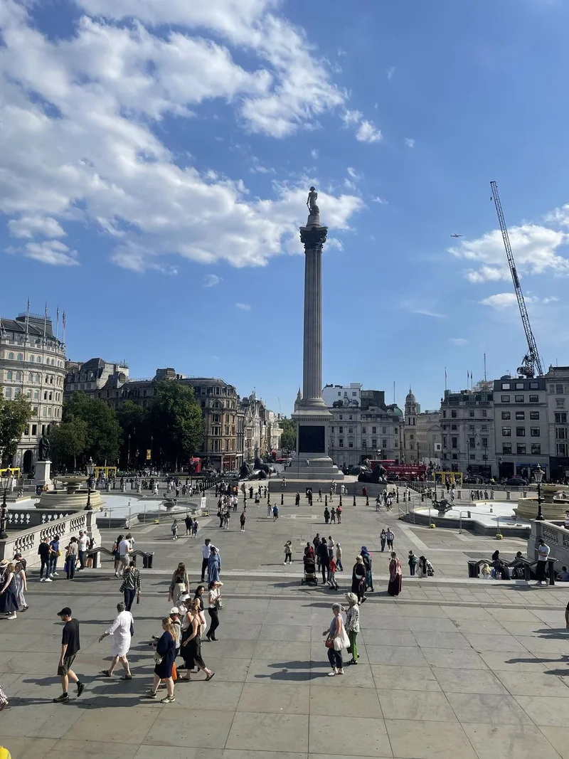 Nelson's Column rising above the paved expanse of Trafalgar Square, with visitors crossing the square under a blue sky.