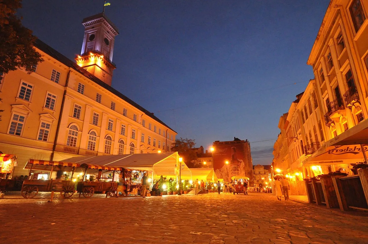 Rynok Square at dusk with the lit Town Hall tower on the left, café stalls and tall townhouses around a cobbled square.