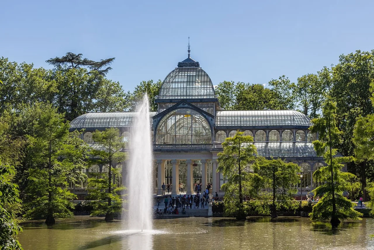 The glass and iron Crystal Palace in El Retiro Park seen across a lake, with a tall fountain jet in the foreground and green trees on both sides.